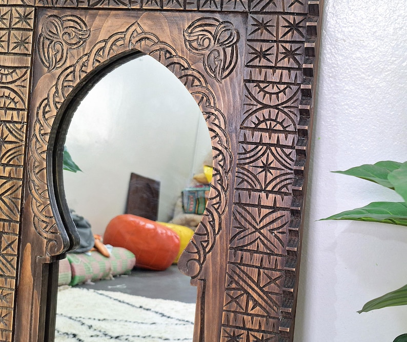 May include: A wooden mirror with an intricately carved frame. The frame features a Moroccan-inspired design with geometric patterns and an arched opening. The mirror is reflecting a room with a rug and furniture.