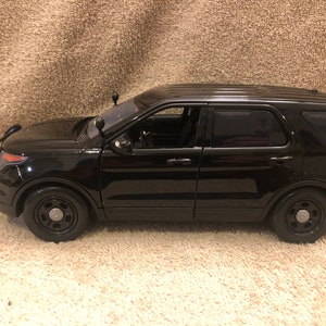 May include: Black toy police car model of a Ford Explorer SUV. The car has a black grill guard and black wheels with silver hubcaps.