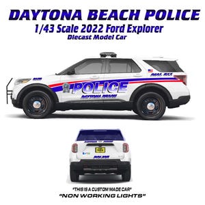 May include: A white and blue diecast model of a Daytona Beach Police Ford Explorer. The model car is 1/43 scale and features the text "POLICE" on the side and "DAYTONA BEACH" on the doors. The text "THIS IS A CUSTOM MADE CAR" and "NON WORKING LIGHTS" is also visible.