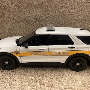 Op de afbeelding: Witte Ford Explorer politieauto met een gele en bruine streep en de tekst "STATE POLICE" aan de zijkant. De auto heeft een zwaailicht op het dak.
