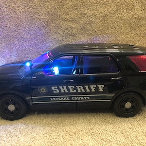 May include: A black toy police SUV with the word "SHERIFF" and "LUZERNE COUNTY" printed on the side. The vehicle has flashing blue and red lights. The headlights are on, and the windows are tinted.
