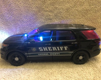 May include: A black toy police SUV with the word "SHERIFF" and "LUZERNE COUNTY" printed on the side. The vehicle has flashing blue and red lights. The headlights are on, and the windows are tinted.