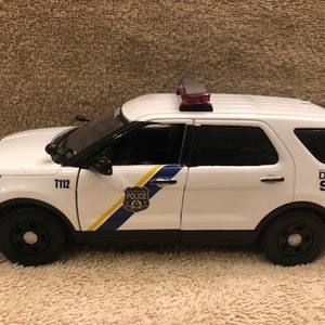 May include: A white and black police SUV toy with the text "T112" and "DIAL 911". The vehicle has a blue and yellow stripe and a police badge on the side. It has black tires and a red and clear light bar on top.