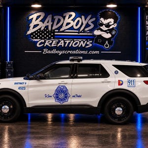 May include: A white police SUV model with black wheels, a push bumper, and blue police decals on the side doors. The vehicle features the text District 5, 4523, 911, and a police badge emblem. The background displays a large wall sign with the text Badboys Creations and a cartoon police officer character.