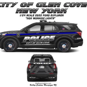 May include: A black and blue 1/24 scale 2022 Ford Explorer police car model. The model has the text "POLICE" and "CITY OF GLEN COVE NEW YORK" on the side. The model also has the text "278" on the side.