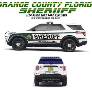 May include: A white 1/24 scale 2022 Ford Explorer model car with green and black Orange County Sheriff decals, working lights and siren. The car has a US flag sticker on the side.