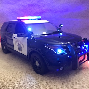 Op de afbeelding: Een zwart-witte speelgoed Ford Explorer politie-SUV met knipperende blauwe en rode lichten. De woorden "HIGHWAY PATROL" zijn zichtbaar op de voordeuren. Het voertuig heeft een voorste pushbar en een kentekenplaat.