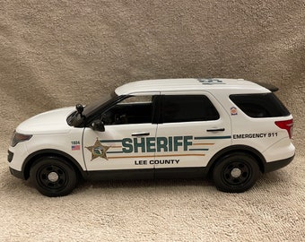 Puede incluir: Coche de policía Ford Explorer blanco con las marcas "Sheriff Lee County" en verde y dorado y "Emergency 911" en la parte trasera. El coche tiene una estrella negra con el número 1824 en el lateral.