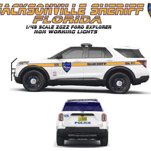May include: A white and yellow 1/45 scale model of a 2022 Ford Explorer Jacksonville Sheriff Florida police car. The model has black wheels and the text "SHERIFF" and "DIAL 911" on the side. The rear of the car is also visible.