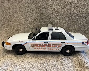 May include: A white toy police car with black wheels and a light bar on top. The car has the words "SHERIFF HARRIS COUNTY" and "NC50" printed on the side. The car has blue and orange stripes.