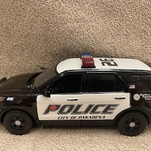 May include: A black and white toy police car with the words "Police City of Pasadena" on the side. The car has a red and blue light on top and the number "26" on the side.