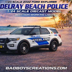 May include: A 1/24 scale diecast model of a 2022 Ford Explorer Delray Beach Police vehicle. The white SUV has blue accents and the text "POLICE" on the side. The model has non-working lights and is parked on a road near a beach.