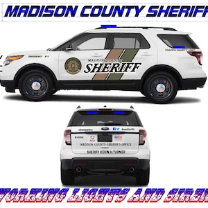 May include: A white Ford Explorer police car with blue and red lights on the roof. The car has a black and tan stripe on the side with the words "Madison County Sheriff" in white letters. The car also has a sticker on the back that says "Madison County Sheriff's Office" and "Sheriff Kevin H. Turner".