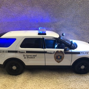 May include: White police car with blue lights, the words "To Serve and to Protect" and the number "21810" on the side. The car has a police badge and the word "Police" on the side.