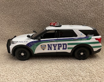 May include: A white NYPD police SUV toy with black wheels and trim. The vehicle has the NYPD logo and the words "POLICE" and "NYPD" printed on the sides. It features red and clear emergency lights on the roof.