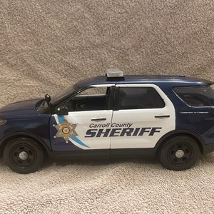 May include: A blue and white toy police car with the words "Carroll County Sheriff" on the side. The car has a light bar on the roof and a star-shaped badge on the door.