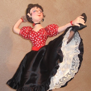May include: A handmade doll wearing a red and white polka dot top, a black skirt with white lace trim, and black shoes. The doll is posed with one leg raised and her arm extended.