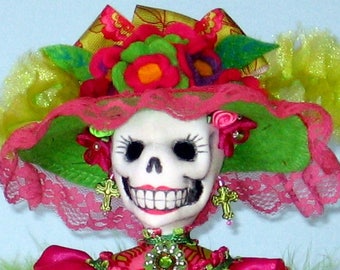 Puede incluir: Una muñeca colorida del Día de Muertos con un sombrero rosa y verde, una cara de calavera blanca y un vestido rosa y verde.