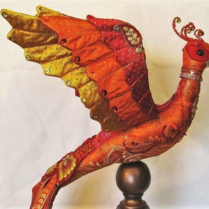 以下が含まれることがあります： 刺繍が施されたオレンジ、赤、金色の手作りの孔雀の置物。孔雀は丸い茶色の台座の上に置かれています。
