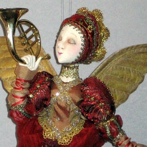 以下が含まれることがあります： 金色の羽根を持つ手作りのお人形の天使。赤と金のドレスと赤と金の冠を着ています。天使は金色のトランペットを持っています。