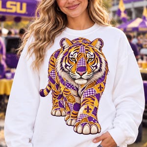 May include: A white long-sleeve sweatshirt featuring a large, central graphic of a tiger. The tiger is composed of a patchwork design in shades of purple, gold, and white, incorporating patterns like gingham, polka dots, and fleur-de-lis symbols.