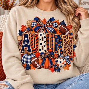 May include: A beige crewneck sweatshirt featuring a large graphic print of the word AUBURN in block letters. The design includes orange and blue pumpkins, footballs, autumn leaves, stars, and bows with leopard, plaid, and polka dot patterns.
