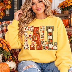 May include: A bright yellow long-sleeve sweatshirt featuring the word FALL in large block letters. The letters are decorated with floral patterns, autumn leaves, and white pumpkins on a green background, creating a seasonal graphic design.