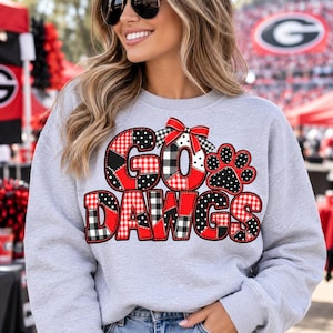 May include: A light gray crewneck sweatshirt featuring a graphic print that reads GO DAWGS in a patchwork style with red, black, and white plaid and polka dot patterns. A red and white plaid bow sits atop the letter O, next to a black paw print.