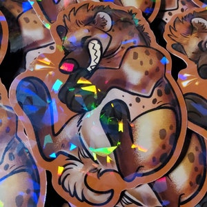 May include: A pile of holographic stickers featuring a cartoon hyena with a toothy grin. The hyena is brown and tan with a white chest and a black nose. The stickers are iridescent and have a rainbow sheen.