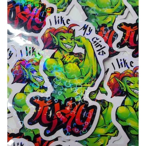 May include: A stack of holographic stickers featuring a green, muscular, cartoon goblin with red hair. The goblin is flexing its bicep and has a toothy grin. The text "I like my girls" is written on the sticker. The stickers have a rainbow holographic finish.