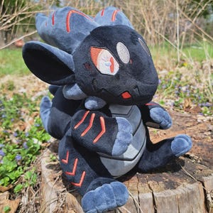 May include: A plush creature toy featuring a black and dark gray body with vibrant red embroidered accents on the head, arms, and legs. The plush has large ears, a light gray belly panel, and detailed eyes with red irises and white highlights.