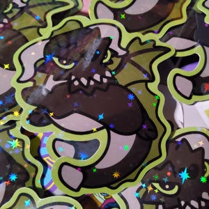 May include: A holographic sticker featuring a black and white cartoon dragon with green and yellow accents. The dragon has a fierce expression and is surrounded by a green outline.