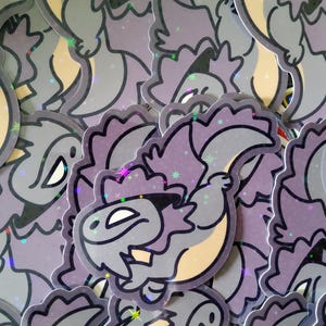 May include: A pile of stickers with a cartoon dinosaur design. The stickers are predominantly grey and purple, with a light tan belly and a holographic finish. The dinosaur has a friendly expression.
