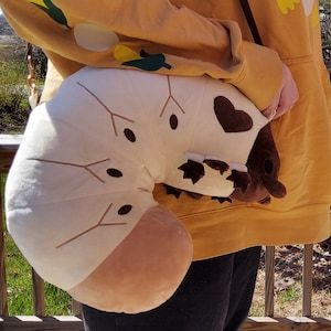 May include: A white and brown plush toy shaped like a grub or caterpillar. The toy has a heart-shaped patch on its back and is being held by a person wearing a yellow hoodie.