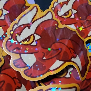 May include: A pile of stickers depicting a red dragon with white details and a yellow border. The dragon has an angry expression and yellow eyes. The stickers have a holographic, glittery finish, with star-shaped sparkles.