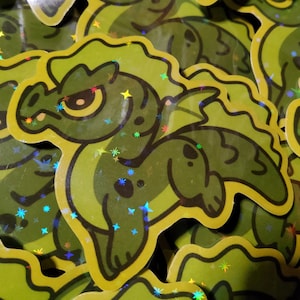 May include: A green holographic sticker of a cartoon dragon with a yellow outline. The dragon has a grumpy expression and is covered in glitter.