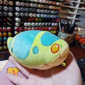 Puede incluir: Un juguete de peluche verde y azul con forma de serpiente con un ojo amarillo. El juguete es suave y esponjoso y sería una gran adición a la colección de juguetes de cualquier niño.