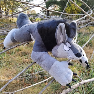May include: A grey and black plush wolf toy with large paws and a detailed face. The toy is lying on twigs and grass, in a playful pose. The wolf has large ears and a fluffy tail. The toy is designed to look like a cartoon wolf.