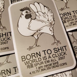 May include: A grey sticker with a black and white illustration of a chicken. The sticker says "Born to Shit World is a Cluck" with the text "鸡蛋 Spur Em All 2007 I am cecal bird 4,10,757,864,350 Dead Cops" underneath.