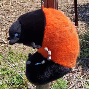 May include: A plush toy shaped like a beetle with a black head and lower body, featuring a bright orange fuzzy midsection. The toy has small grey feet, a blue rectangular accent on the lower section, and a silver metal clip attached to the top.