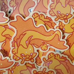 May include: A pile of stickers depicting a cartoon creature in shades of orange and yellow, with a long, flowing tail. The stickers have a glossy finish and a white border. The creature has a smiling face and star-shaped details.