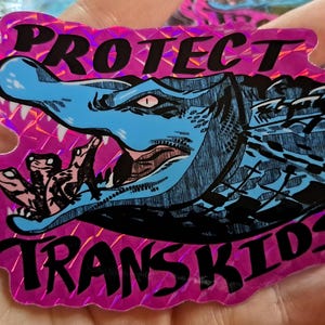 May include: A holographic sticker with a pink background and the words "PROTECT TRANS KIDS" in pink. The sticker features a blue alligator with an open mouth, and several smaller alligators inside.