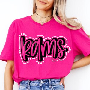 May include: A bright pink t-shirt with the word "RAMS" in a black, cursive font, outlined in white and dotted with white circles. The shirt is made of a soft material and is a crew neck style.