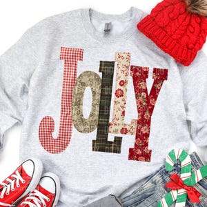 May include: A light gray sweatshirt with the word "JOY" appliqued in various patterned fabrics, including red gingham, floral, and plaid. A red knit beanie and red sneakers are also in the image. The theme is festive and holiday-oriented.