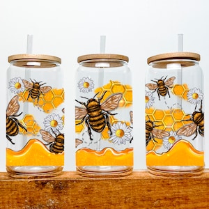 May include: Three clear glass tumblers with bamboo lids and straws. Each tumbler features a design of bees, honeycombs, and daisies in yellow, orange, and white.
