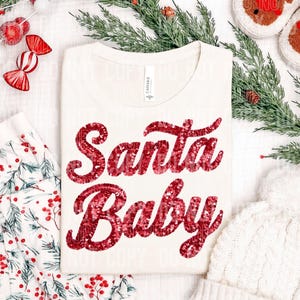 May include: A cream-colored t-shirt featuring the text Santa Baby written in shiny red sequins across the chest. The shirt has a round neckline and a white tag at the collar. The design is centered on the front of the garment.