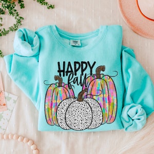May include: A light blue sweatshirt with the words "HAPPY FALL" in black. The sweatshirt features three pumpkins, two with colorful plaid patterns and one with a black and white polka dot design. The sleeves are folded.