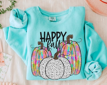 May include: A light blue sweatshirt with the words "HAPPY FALL" in black. The sweatshirt features three pumpkins, two with colorful plaid patterns and one with a black and white polka dot design. The sleeves are folded.