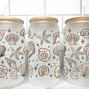 May include: Three frosted glass tumblers with wooden lids and straws. Each tumbler is decorated with a pattern of seashells, starfish, and sparkling accents in beige and black. The tumblers are designed for cold beverages.