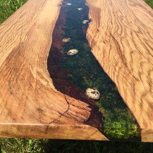 Pode incluir: Uma mesa de centro de madeira com um design de borda viva. O tampo da mesa apresenta um rio de resina com um esquema de cores verde e marrom e ovos de codorniz salpicados de branco embutidos na resina.